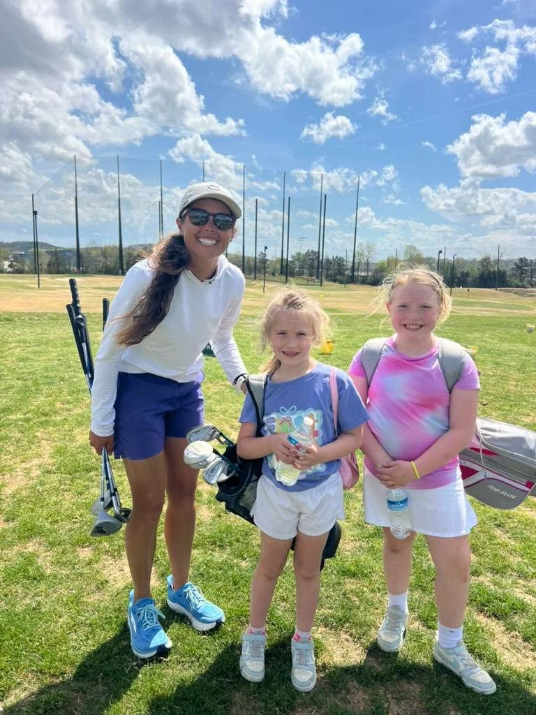 A woman and two young girls standing on a golf course, holding golf clubs and golf balls, under a partly cloudy sky.