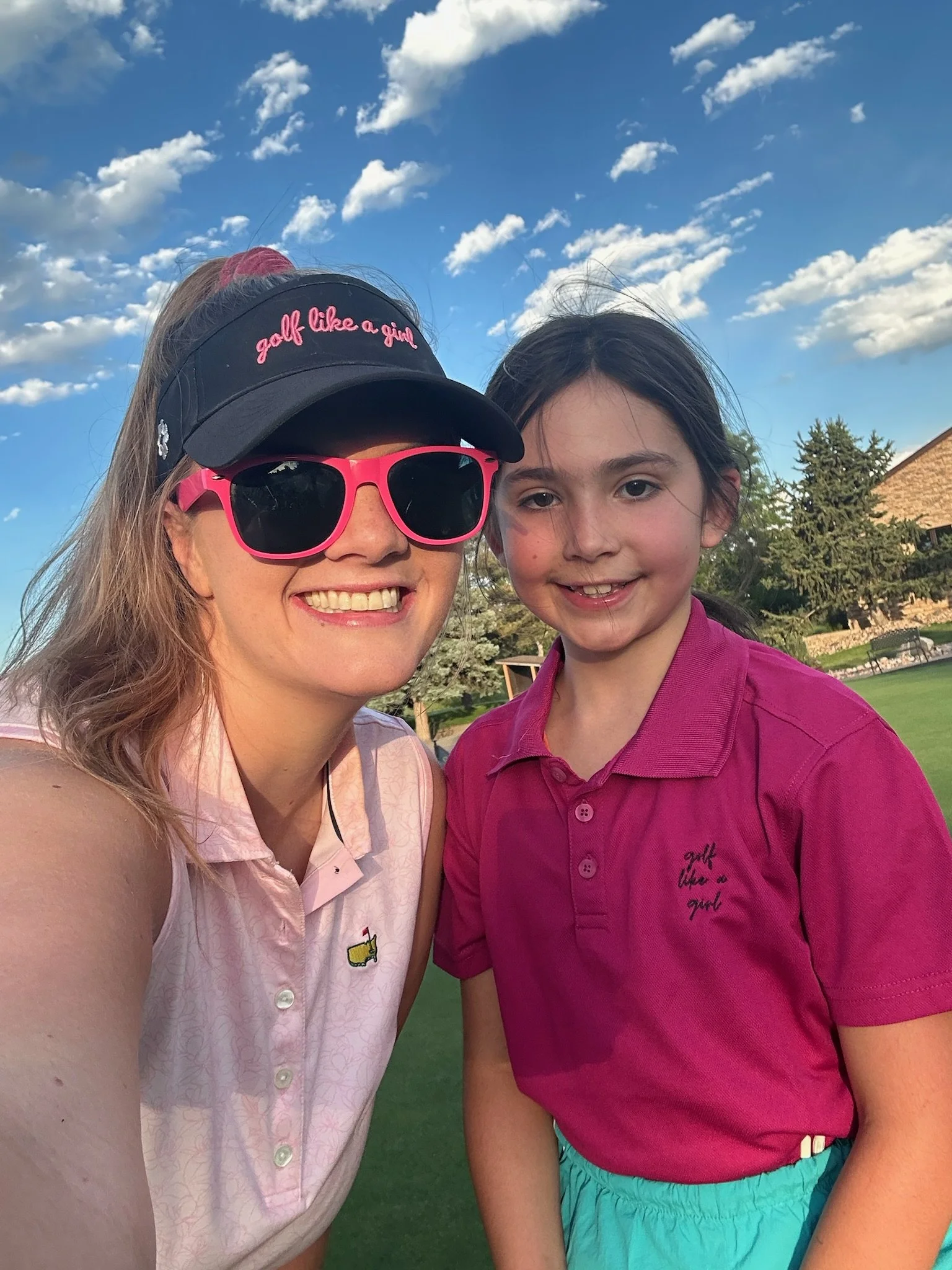 A woman and a girl taking a selfie outdoors on a sunny day, with blue sky and scattered clouds in the background. The woman is wearing a black visor with pink embroidery that says "golf like a girl" and pink sunglasses. The girl is wearing a pink pol