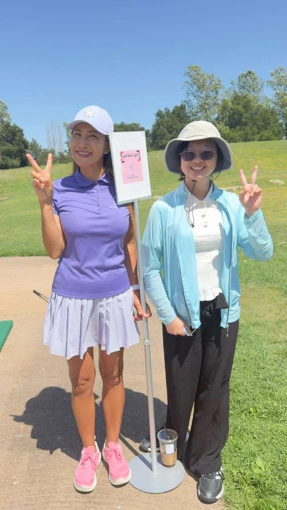Two women showing peace signs at a golf course. One is holding a sign, and the other is wearing sunglasses and a large sun hat.