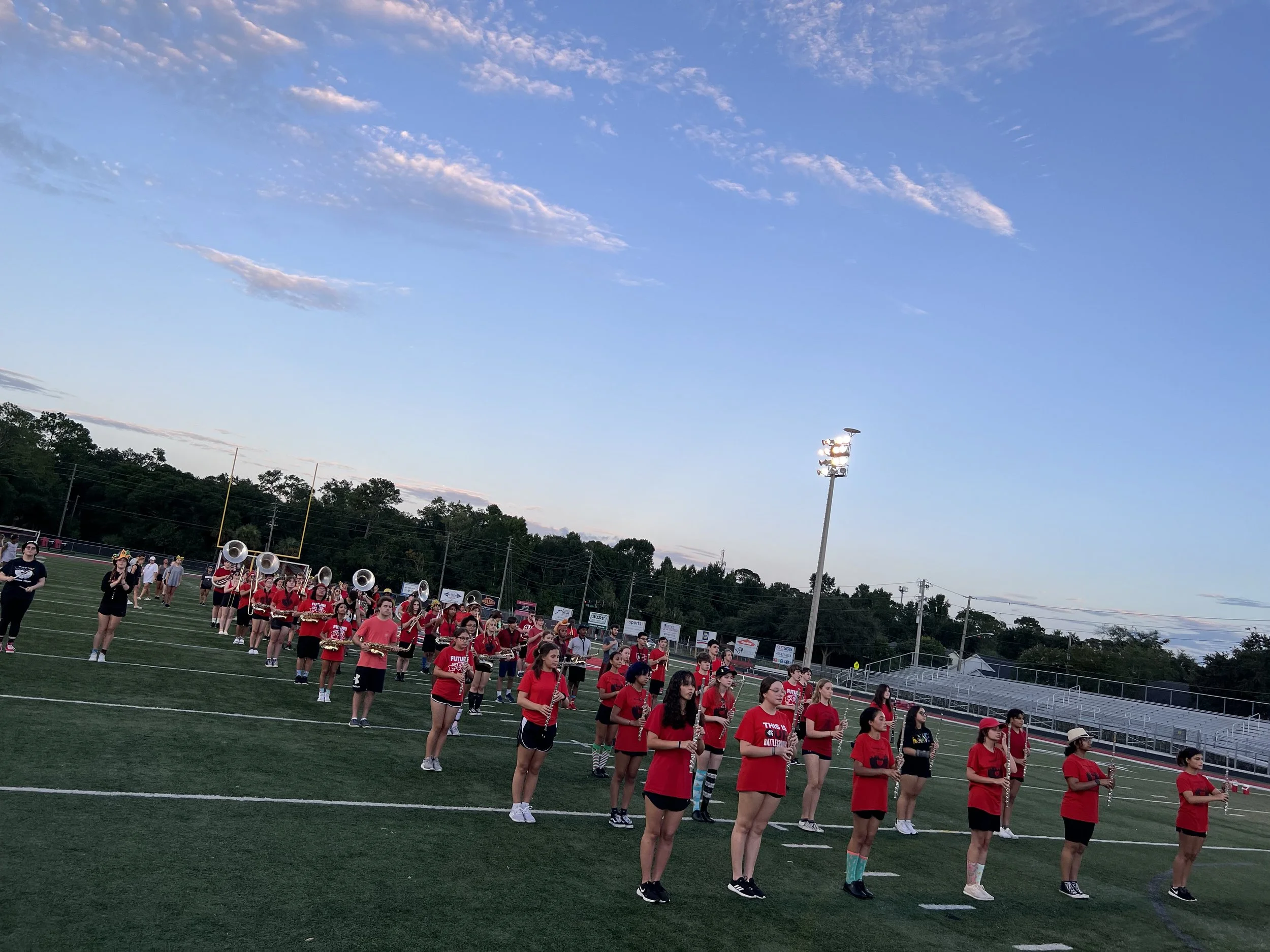 A marching band practicing on a football field during sunset, with band members in red shirts holding instruments, and a clear sky with few clouds overhead.