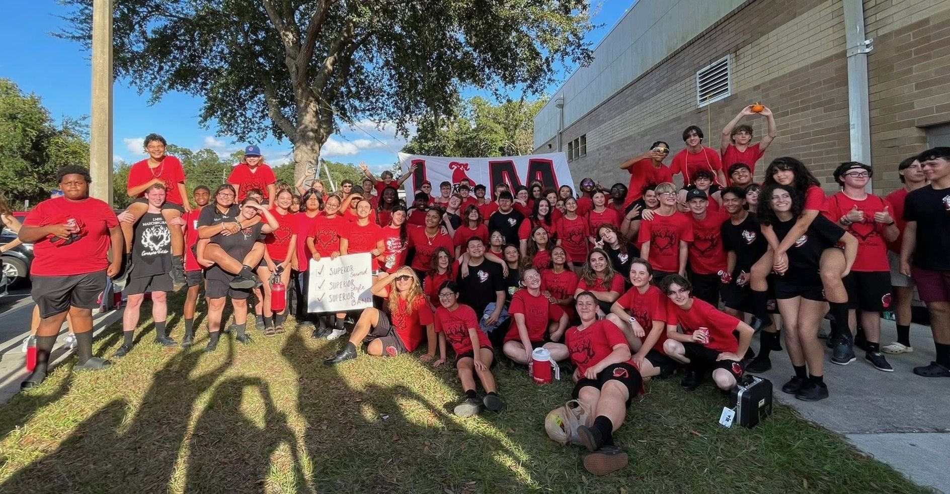 Large group of students wearing red and black shirts, some holding signs and a pumpkin, gathered outside a school building for a celebration or event, with a tree and blue sky in the background.