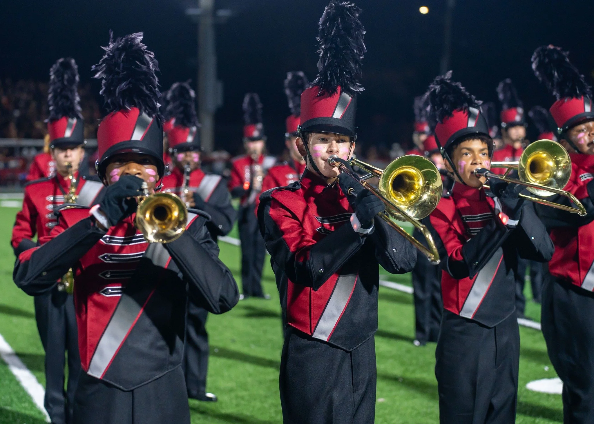 High school marching band performing on a football field at night, with band members wearing red and black uniforms with tall feathered hats, playing brass instruments.