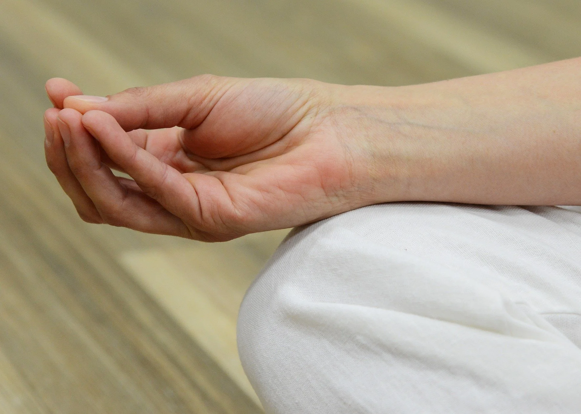 A person's hand is resting on their lap, with the palm facing upward and the fingers gently curved. The person is wearing white pants, and the background appears to be a light, wooden floor.
