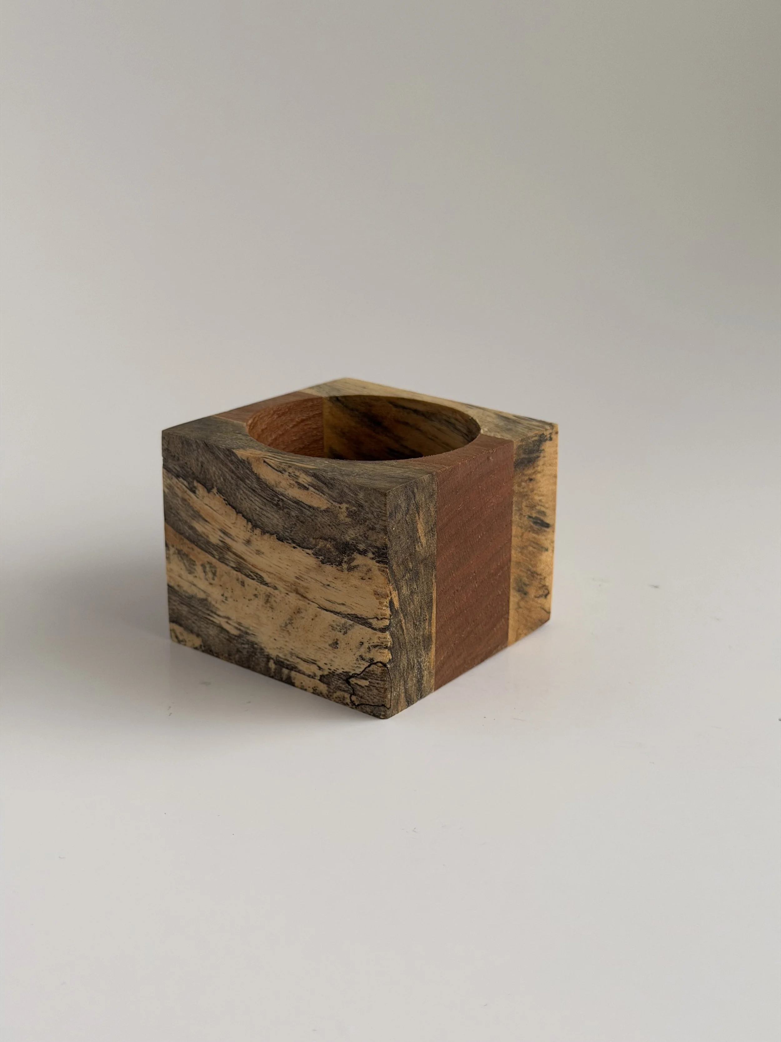 A small wooden cube with a circular hole in the top, displaying varied wood grain patterns and colors on each side, set against a plain white background.