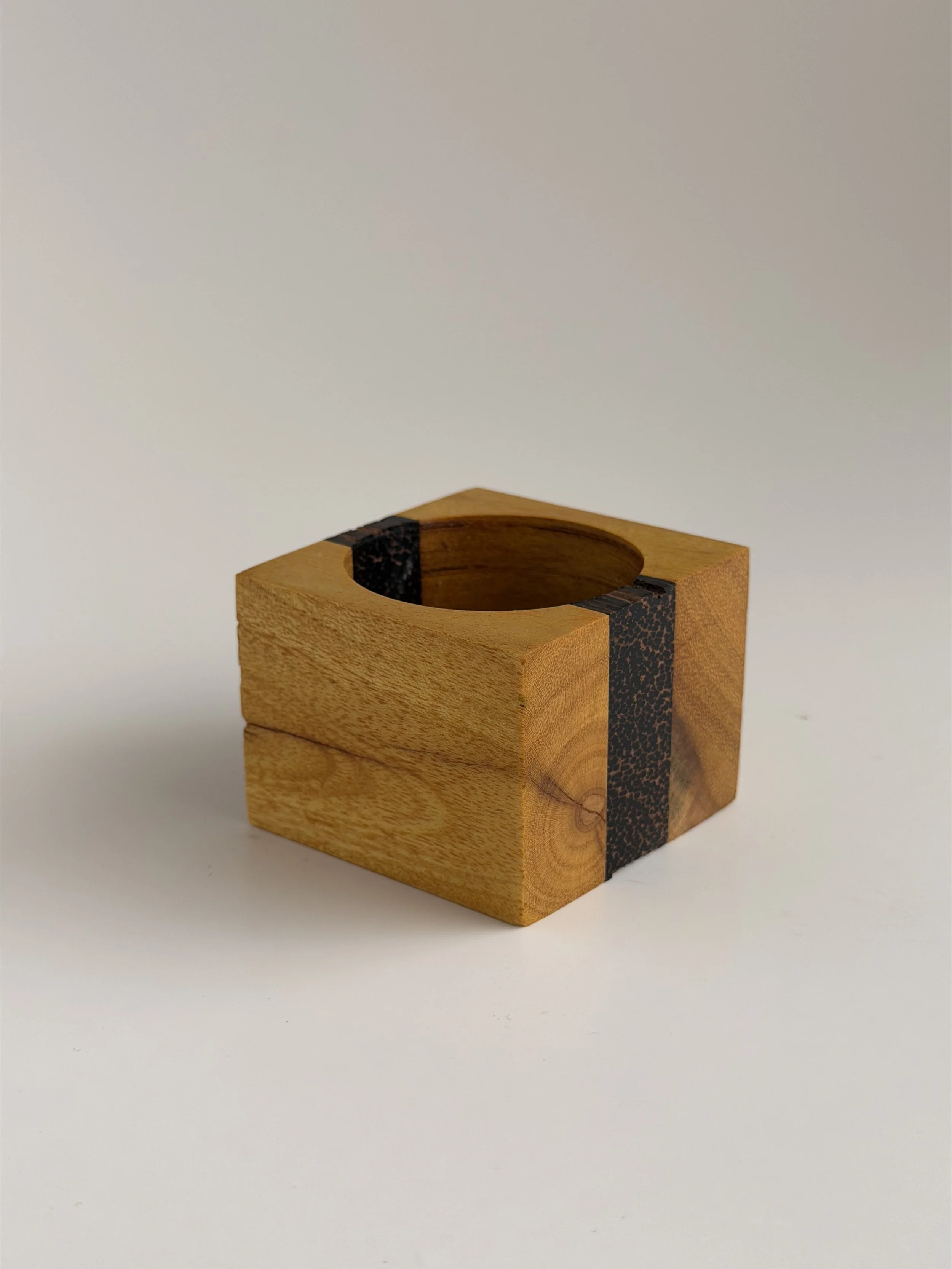 A wooden tissue box holder with a square shape, featuring a round opening on top and a black decorative stripe on one side, placed on a plain white surface.