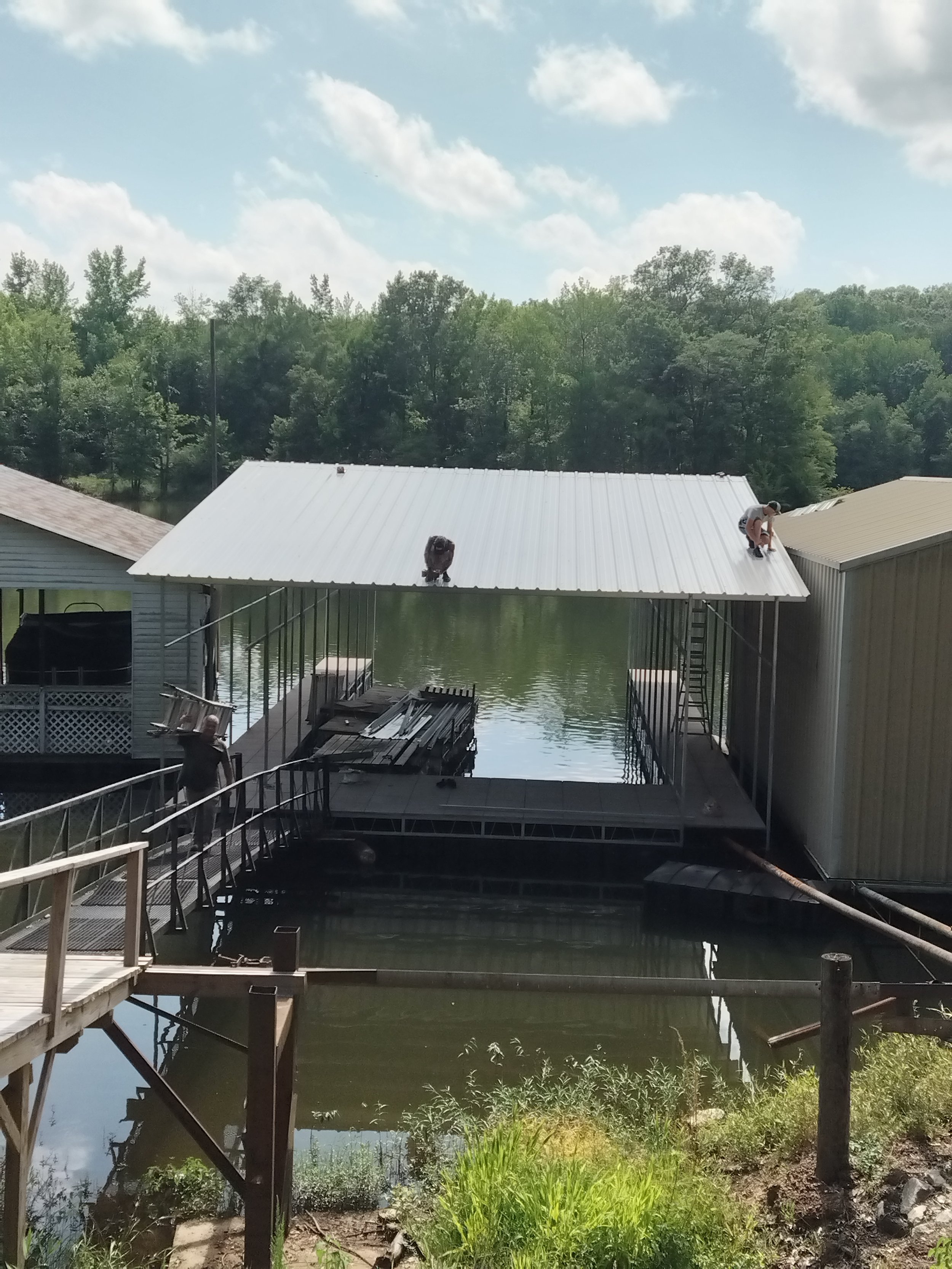 Two people are installing or repairing a white metal roof on a dockside building over a body of water, with construction tools visible and grassy plants in the foreground.