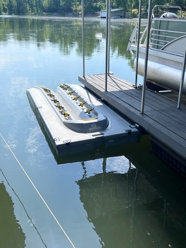 A docked pontoon boat with a folded water slide attached, floating on a calm lake next to a wooden dock, with lush green trees in the background.