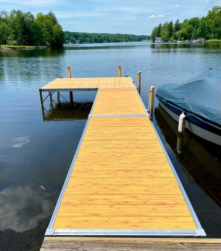 A wooden dock extending into a calm river, with a covered boat moored on the right side and lush green trees lining the riverbanks on both sides under a partly cloudy sky.