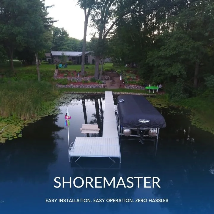 A lakeside Shoremaster dock with a boat cover, a floating dock with a flag, and a garden with a house in the background.