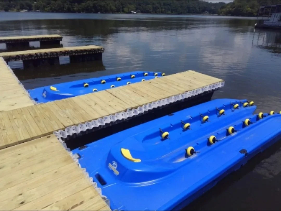 Two blue jet ski floating dock sections with built-in slip-resistant ramps, floating on a calm lake, with a wooden dock section connecting them, and in the background, a ship is docked on the shoreline.