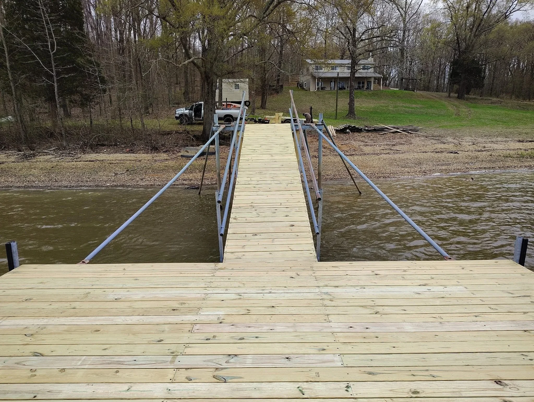 Newly built wooden dock extending over a calm body of water with a metal railing on each side, leading towards the shore where trees, a house, and a parked truck are visible.