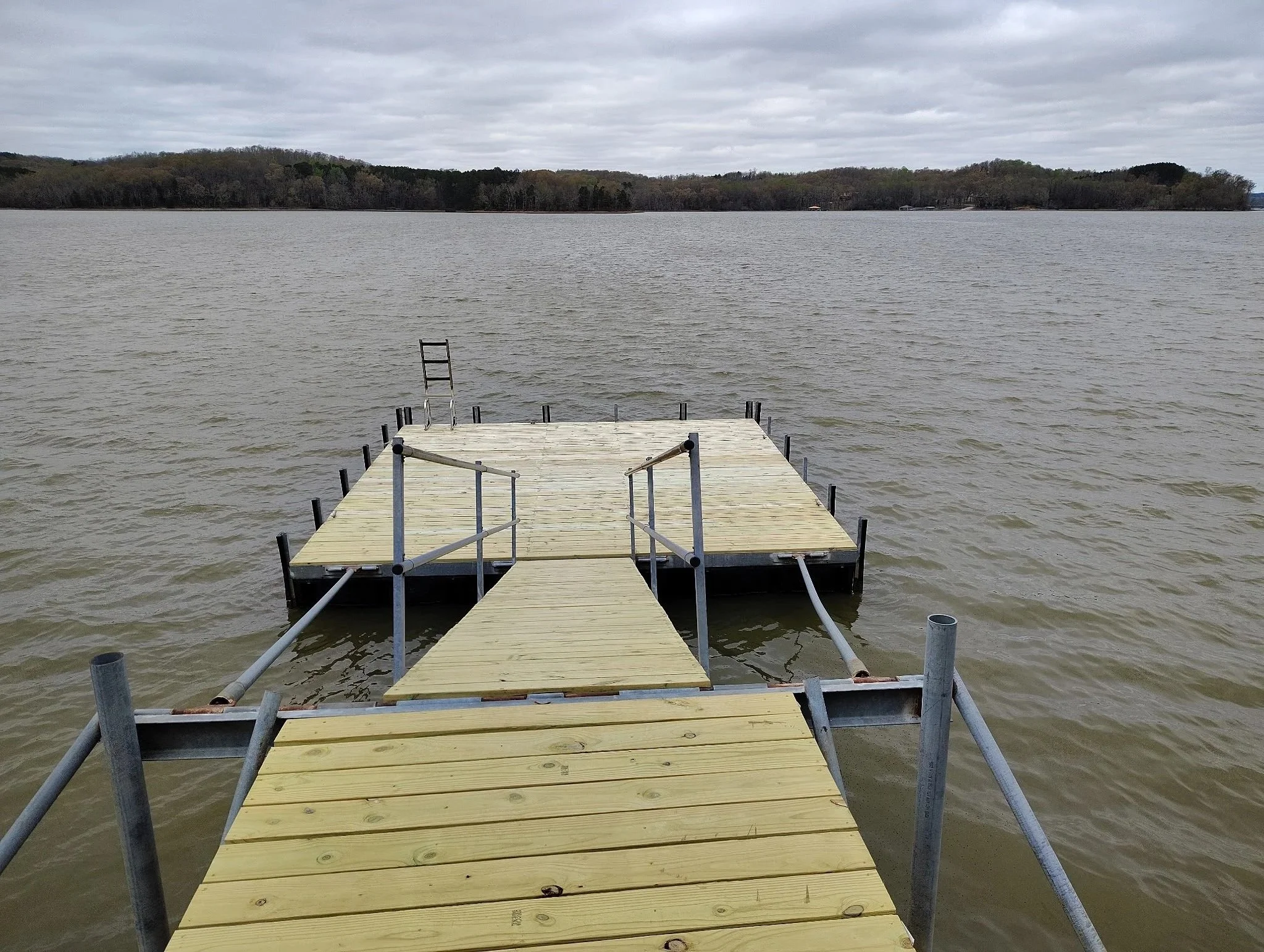 A wooden dock extending into a lake with overcast sky and distant tree-lined shoreline.