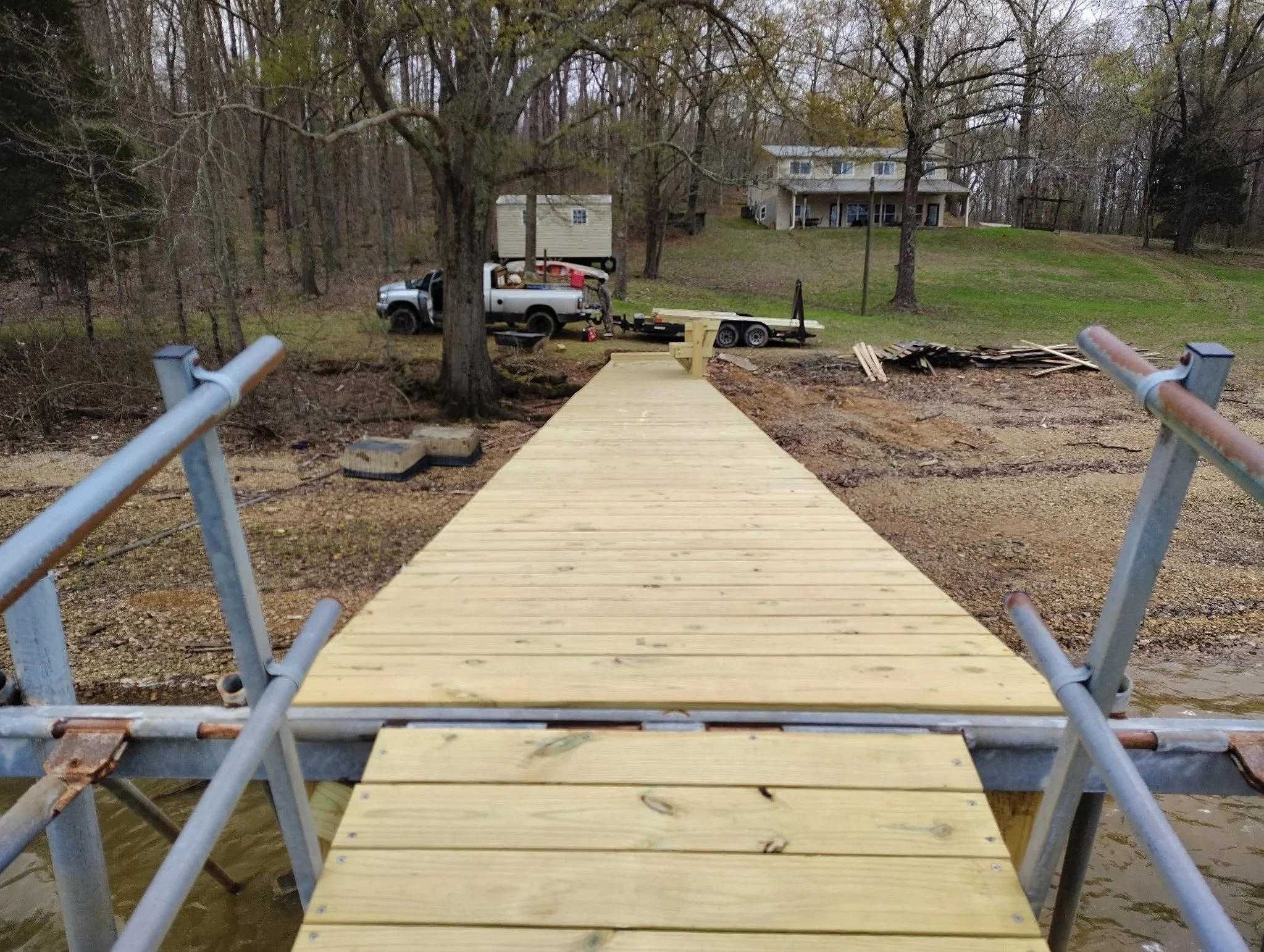 A newly constructed wooden dock extends into a lake, with a metal railing on either side, leading up to a grassy backyard with large trees, a house, a pickup truck, and construction tools and materials in the background.