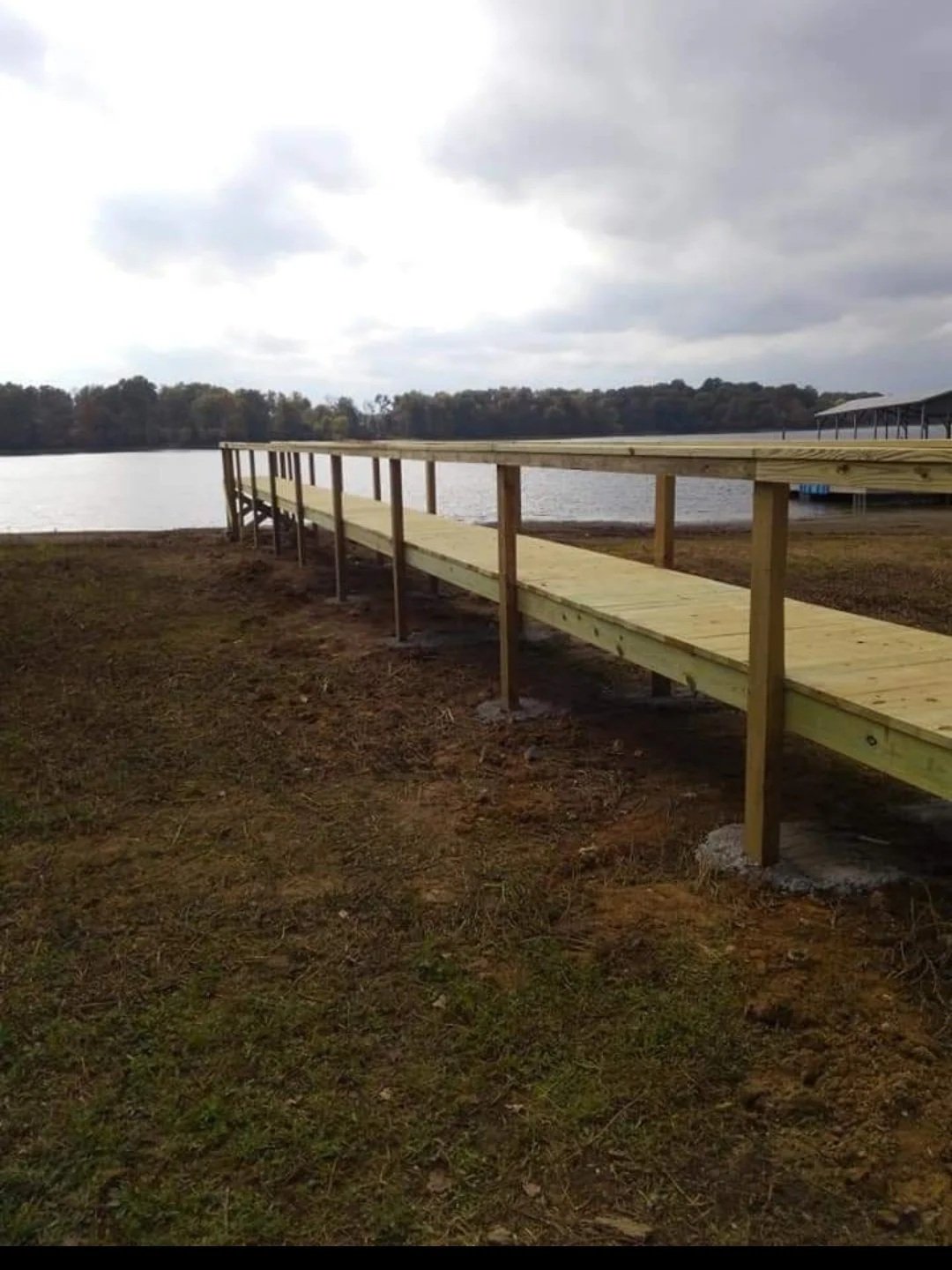 A newly built wooden dock walkway extending toward a body of water under cloudy sky, with trees in the background.