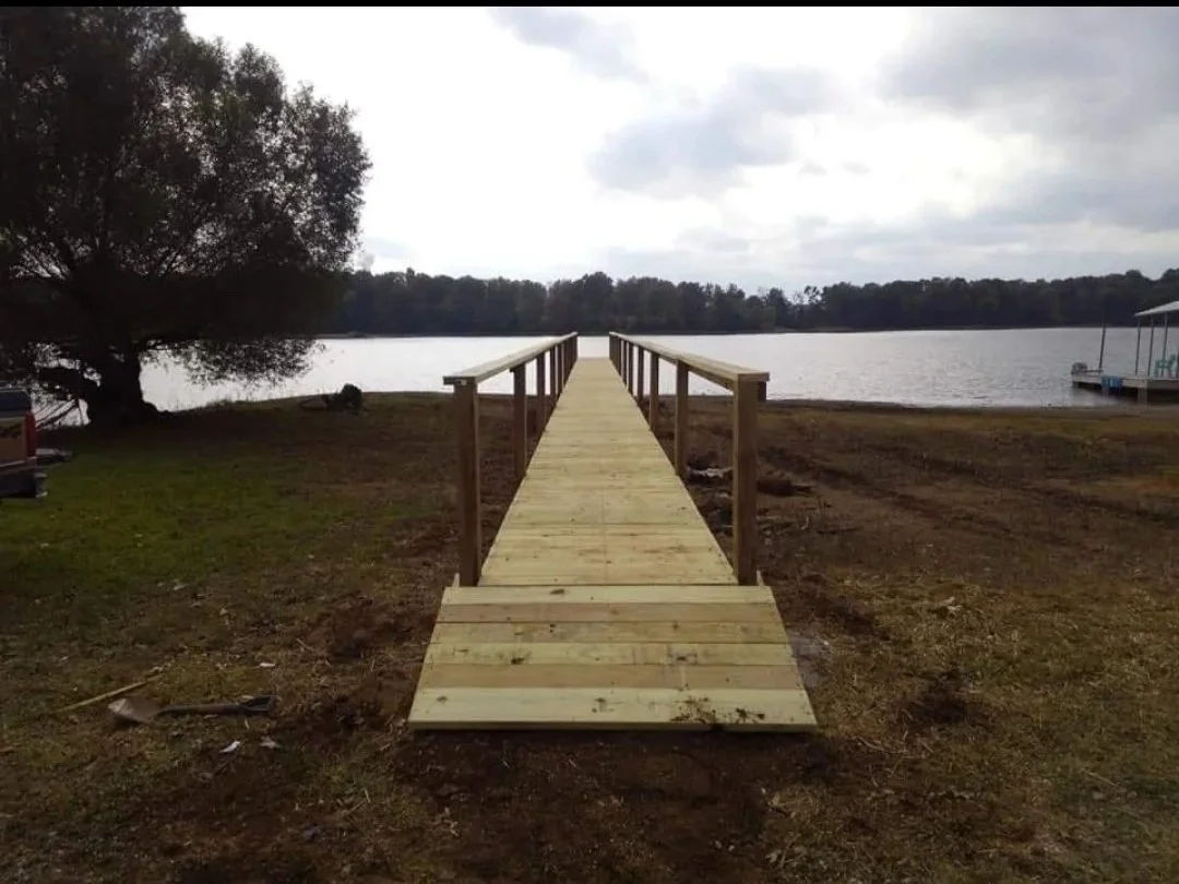 A newly built wooden dock extends over a body of water, with a ramp at the entrance, bordered by a railing on both sides, leading to a tree-lined shoreline with overcast skies overhead.