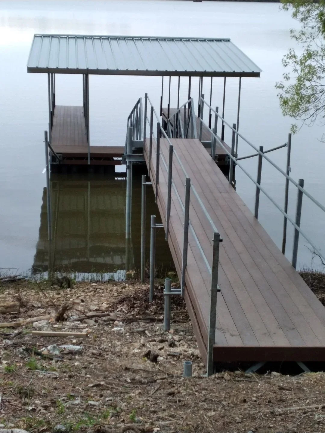 A wooden dock with a metal roof extending over the water, connected by a ramp, with a railing along the sides, situated on a lakeshore with a dirt and grassy bank and some trees.