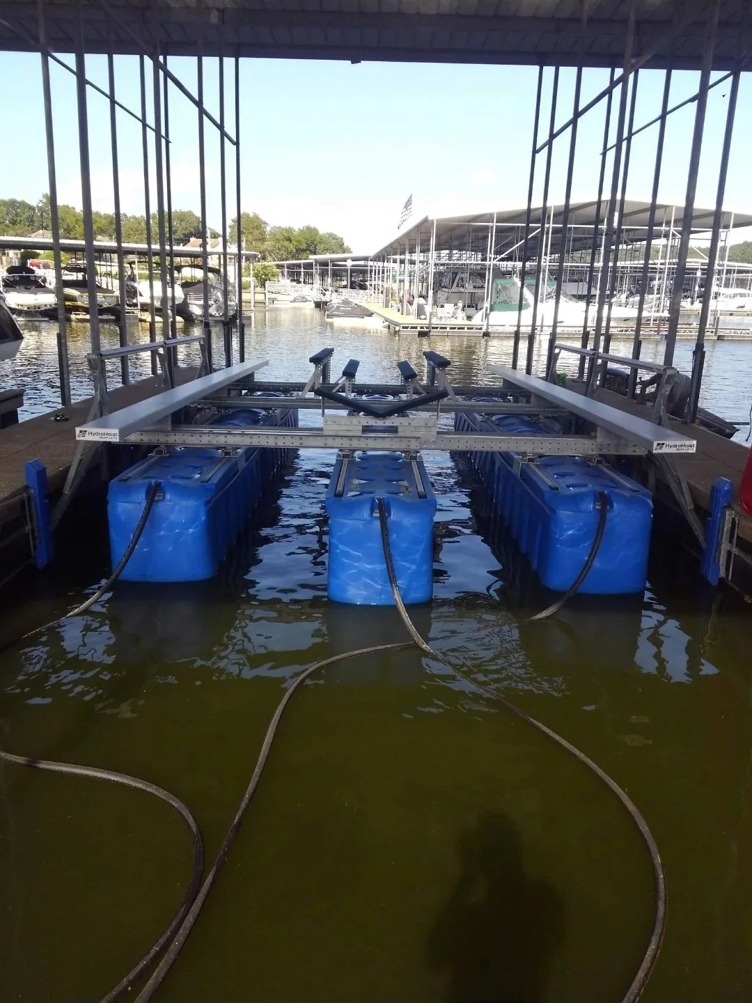 Dock with a boat lift fitted with Hydrolift tanks and a mechanical arm, at a marina with boats and floating docks in the background.