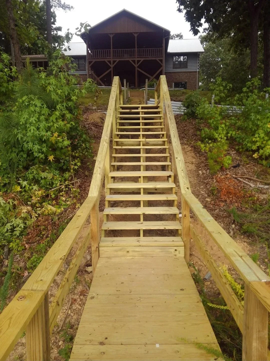 Lake dock connected to wooden staircase leading up a hill towards a house with a balcony and metal roof, surrounded by trees and lush green foliage.