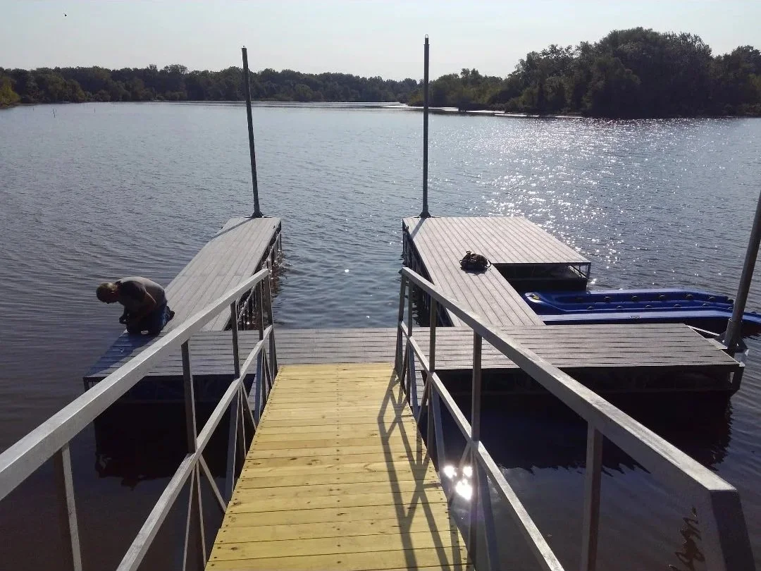 View of a wooden dock extending into a lake with two floating platforms on either side.  Trees are visible in the background under a clear sky.