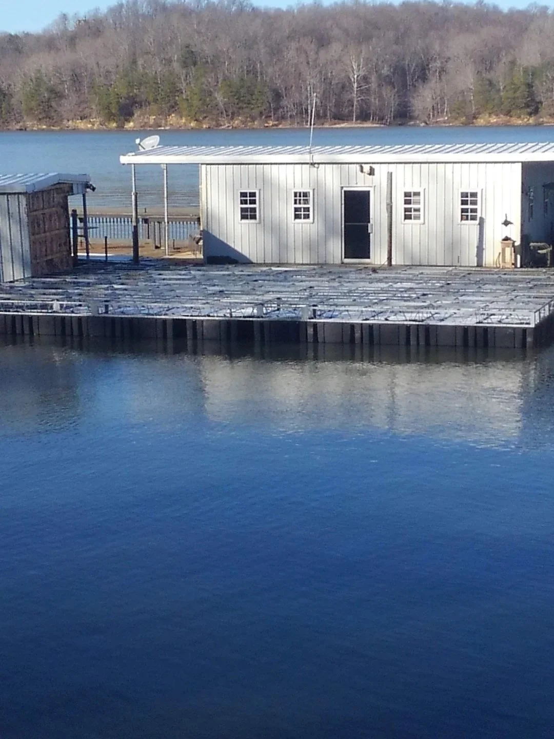 A waterfront scene with a building next to a body of water, with trees on a hill in the background. The building has a metal roof, multiple windows, and a satellite dish, and there is a wooden dock in the water.