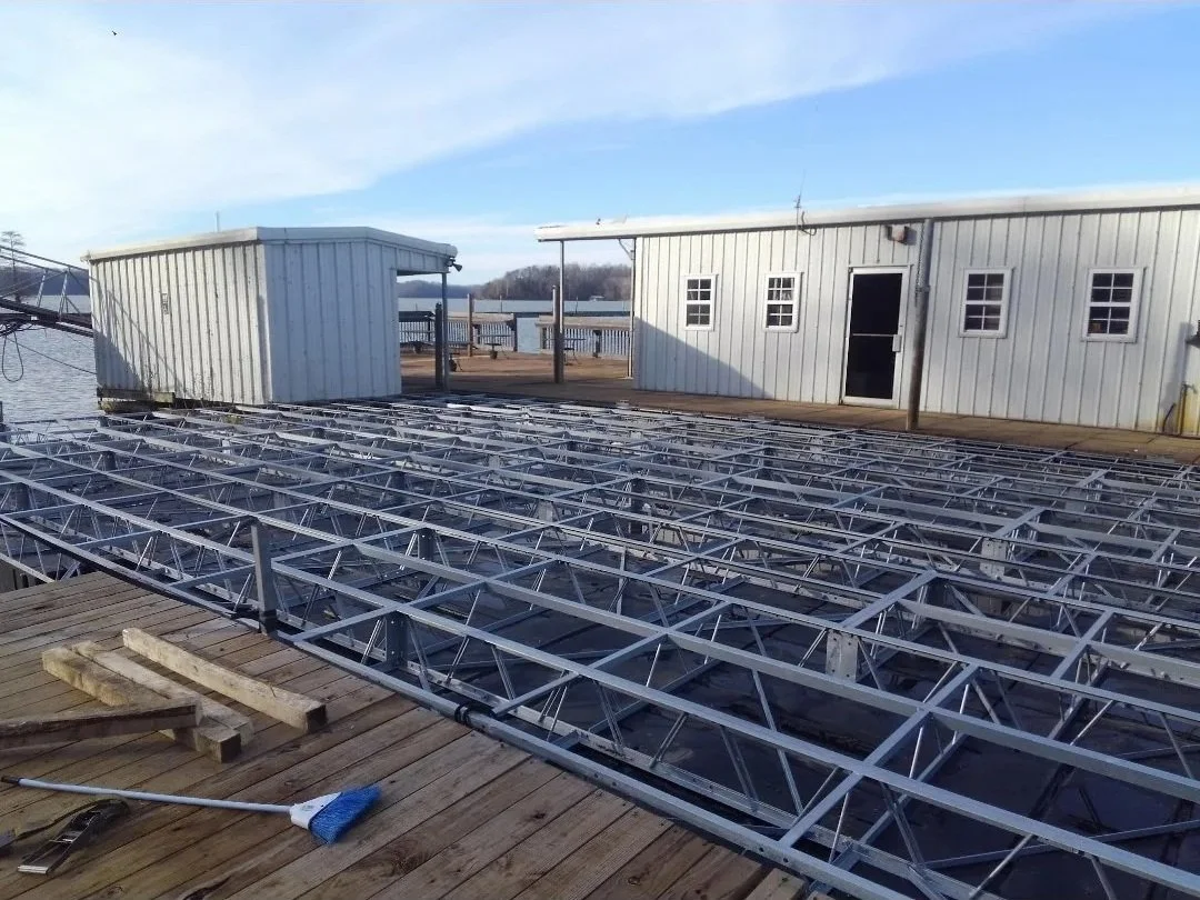 Metal framework being assembled on a wooden dock near the water, with a small white building and clear blue sky in the background.