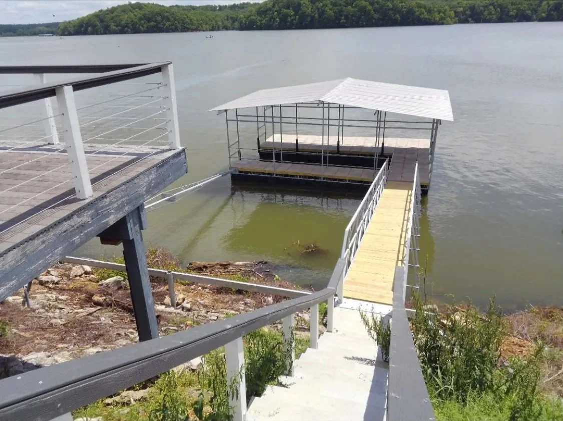 A dock with stairs leading down to a covered floating platform on a lake, surrounded by green trees.