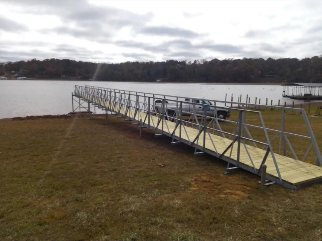A metal and wooden dock extending into a body of water on a cloudy day with a car parked on the dock and grassy land in the foreground.