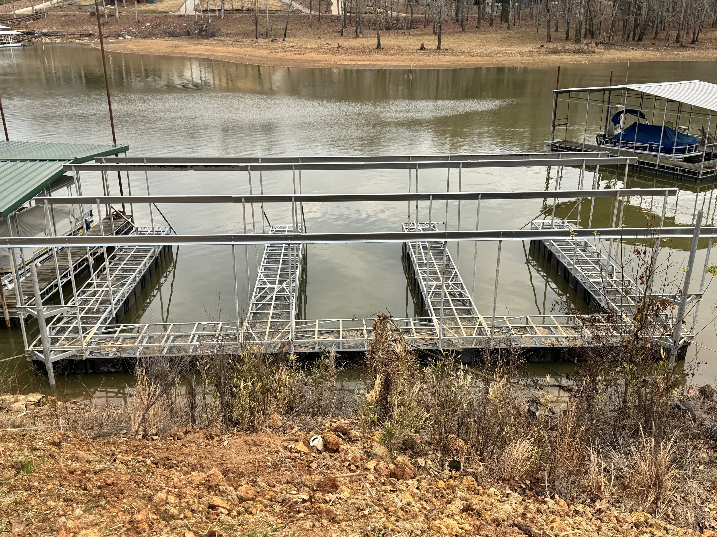 A marina with empty boat docks and a covered boat slip on the water. The water is calm, and the opposite shore has a grassy area with trees.