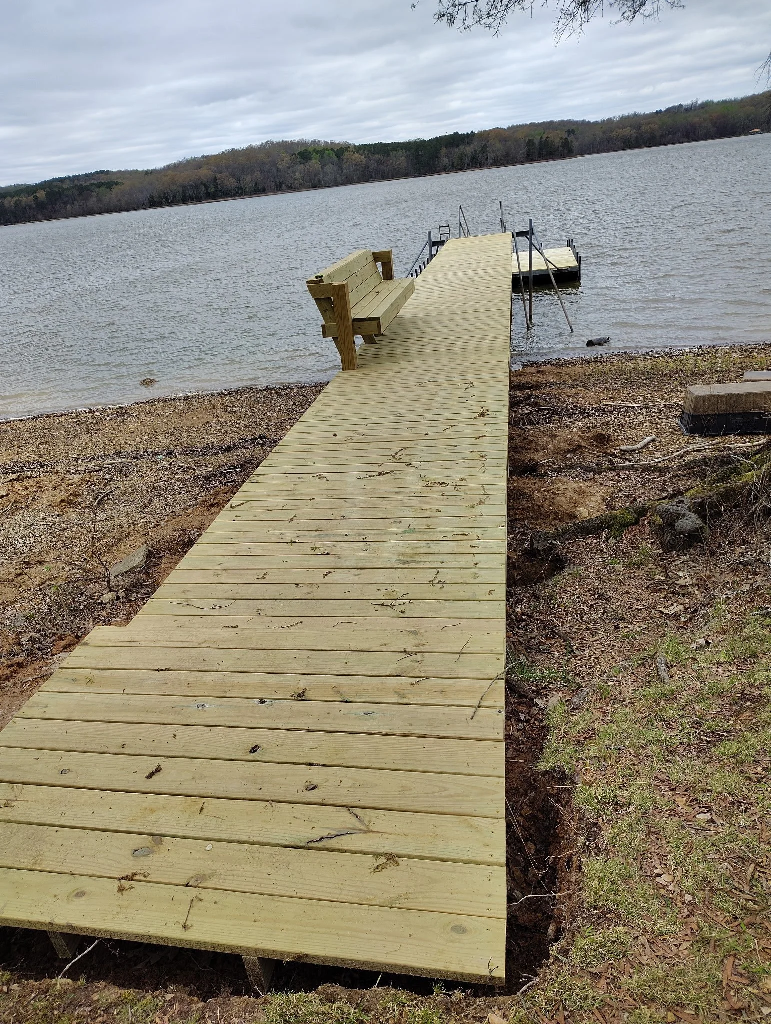 Wooden dock extending into a lake with a bench on the left side, metal handrails on the right, and a ladder at the end of the dock in a cloudy outdoor setting.