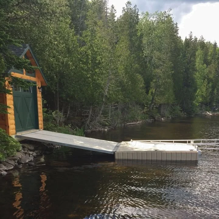 A shoreline with a small, wooden boathouse and a fold-down boat dock extending into a body of water, surrounded by green trees.