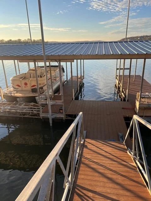 A wooden dock extending into a calm body of water with a boat covered under a metal roof on the left side and a smaller dock on the right. The sky is partly cloudy with the sun shining.