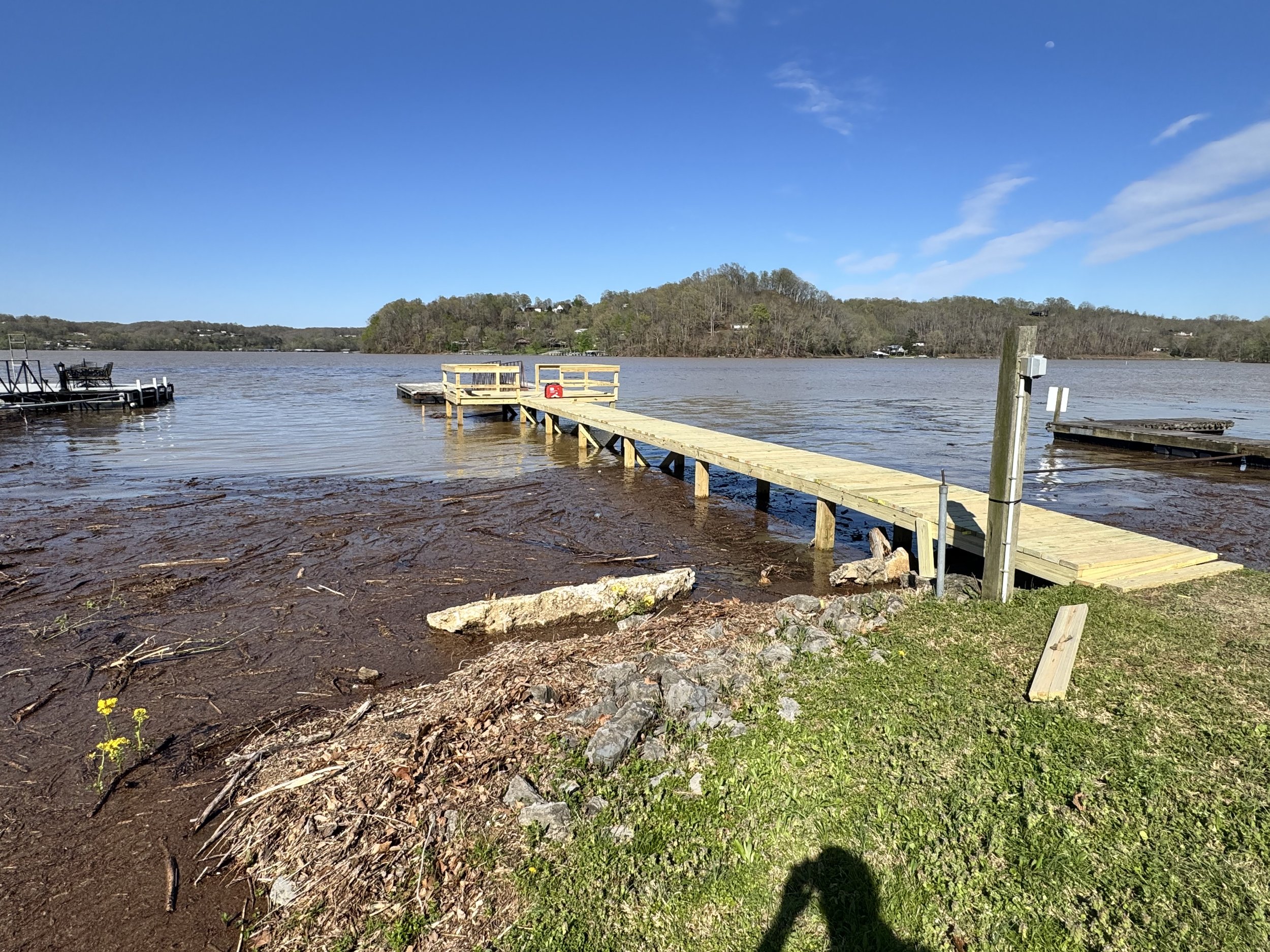 A wooden dock extending into a lake with surrounding trees, blue sky, and some clouds, with a shadow of a person taking the photo visible in the foreground.