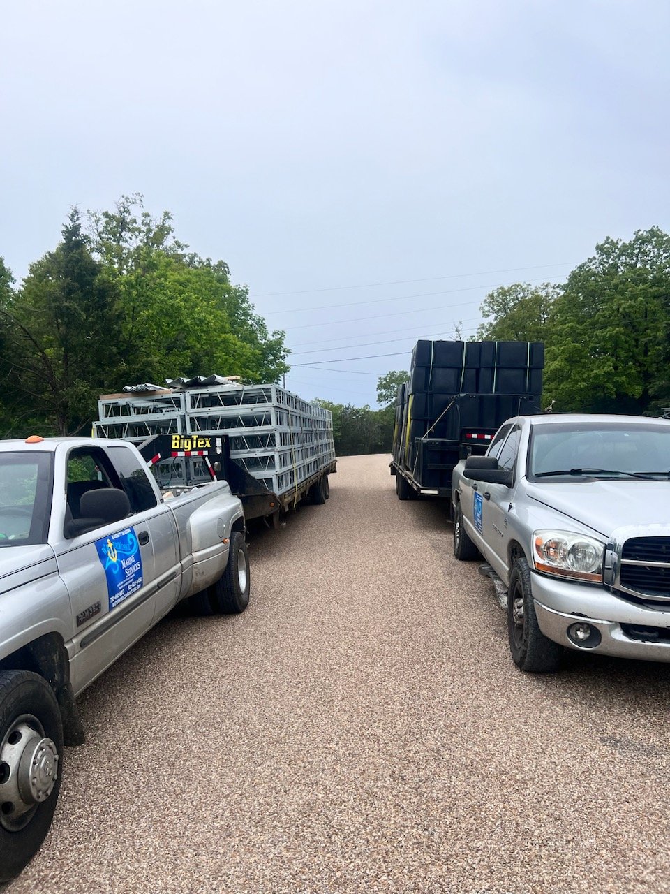 Two pickup trucks parked on a gravel road, loaded with construction materials and black water tanks, surrounded by green trees under a cloudy sky.
