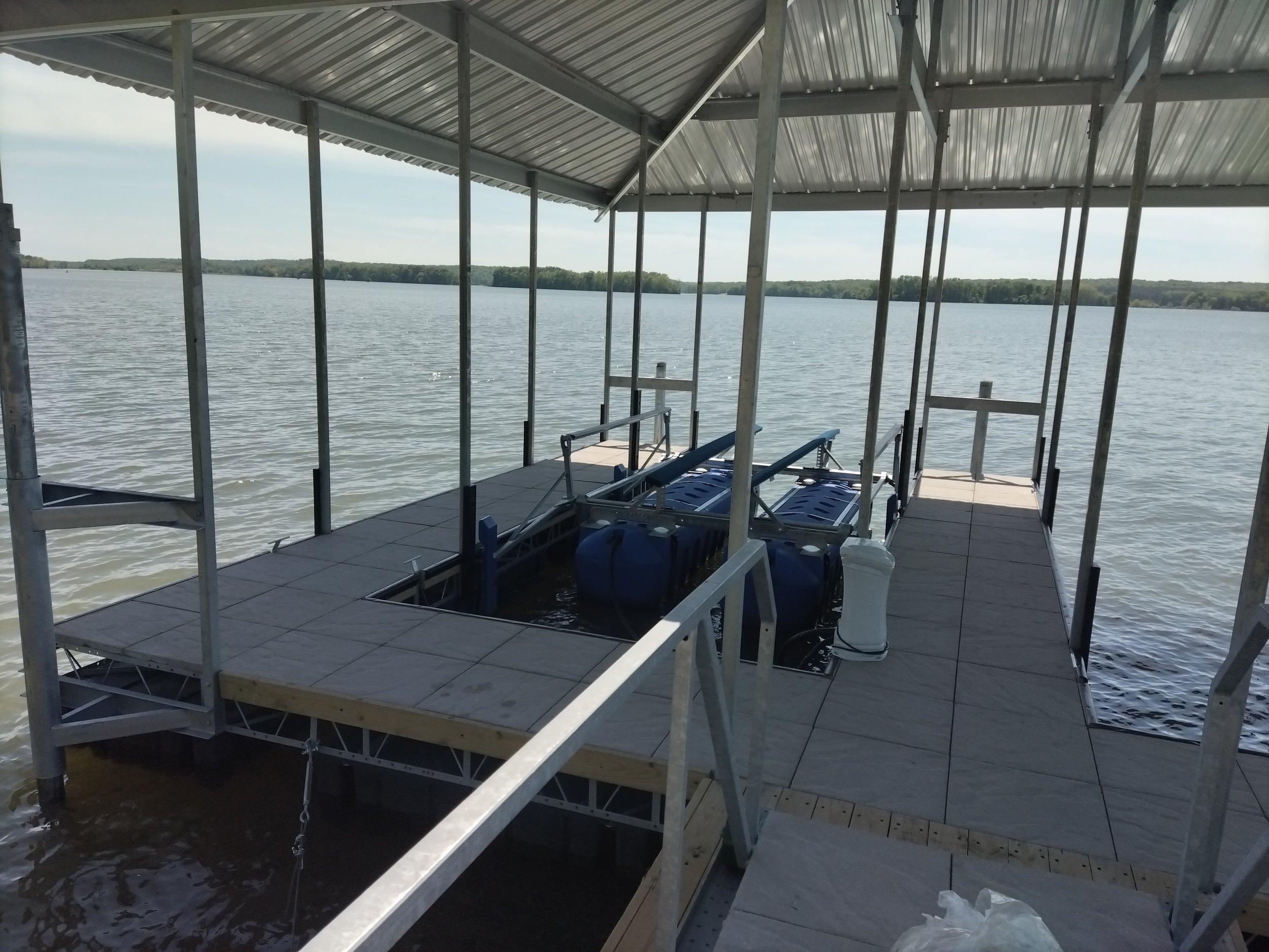 A dock with a metal roof and tile flooring extends over a lake, with some submerged equipment and tanks visible underneath the dock.