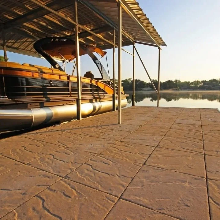 A pontoon boat docked under a metal carport on a stone patio near a calm lake at sunset.