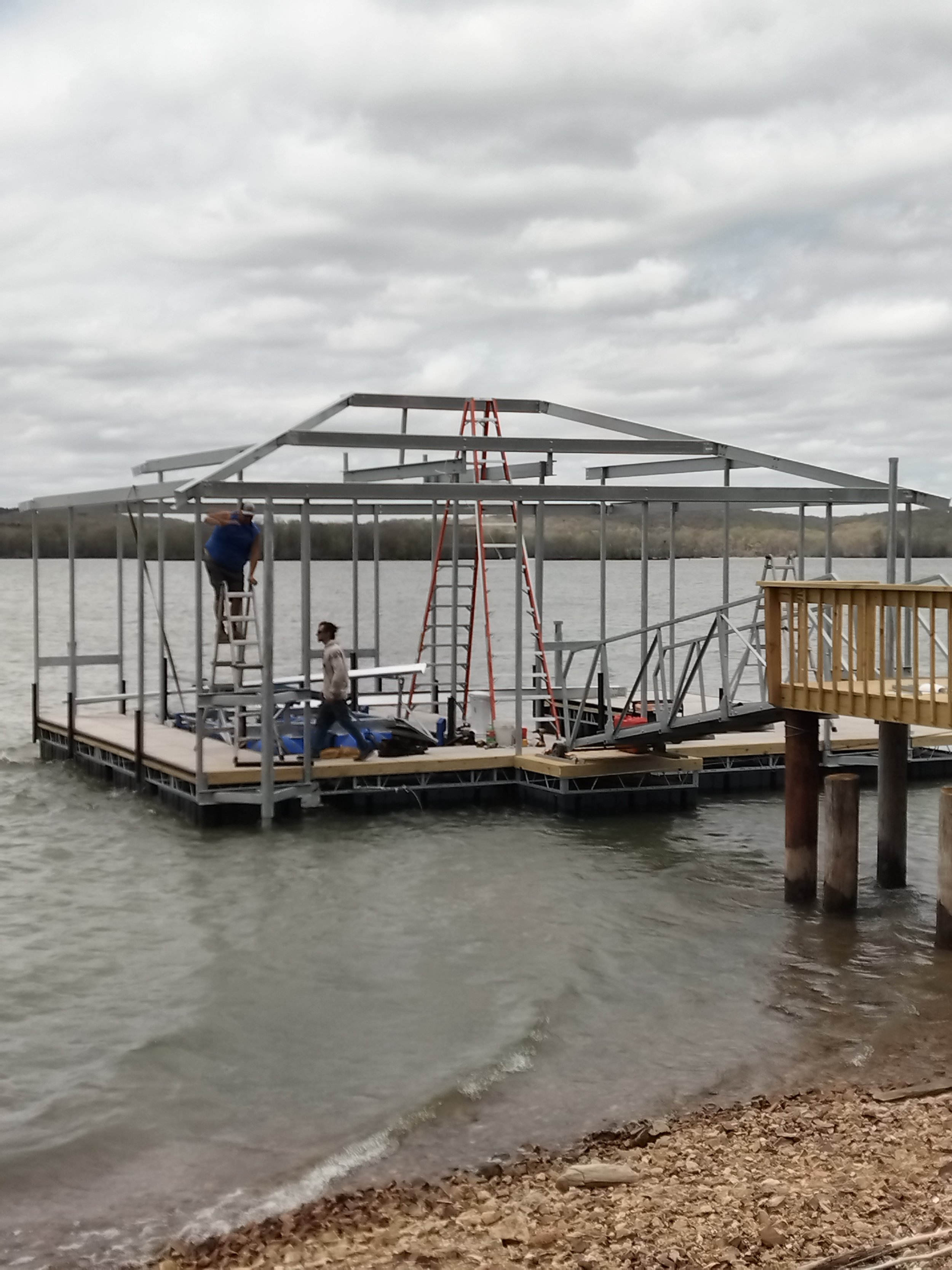 Men working on the frame of a dock under construction on a cloudy day, with water and land in the background.