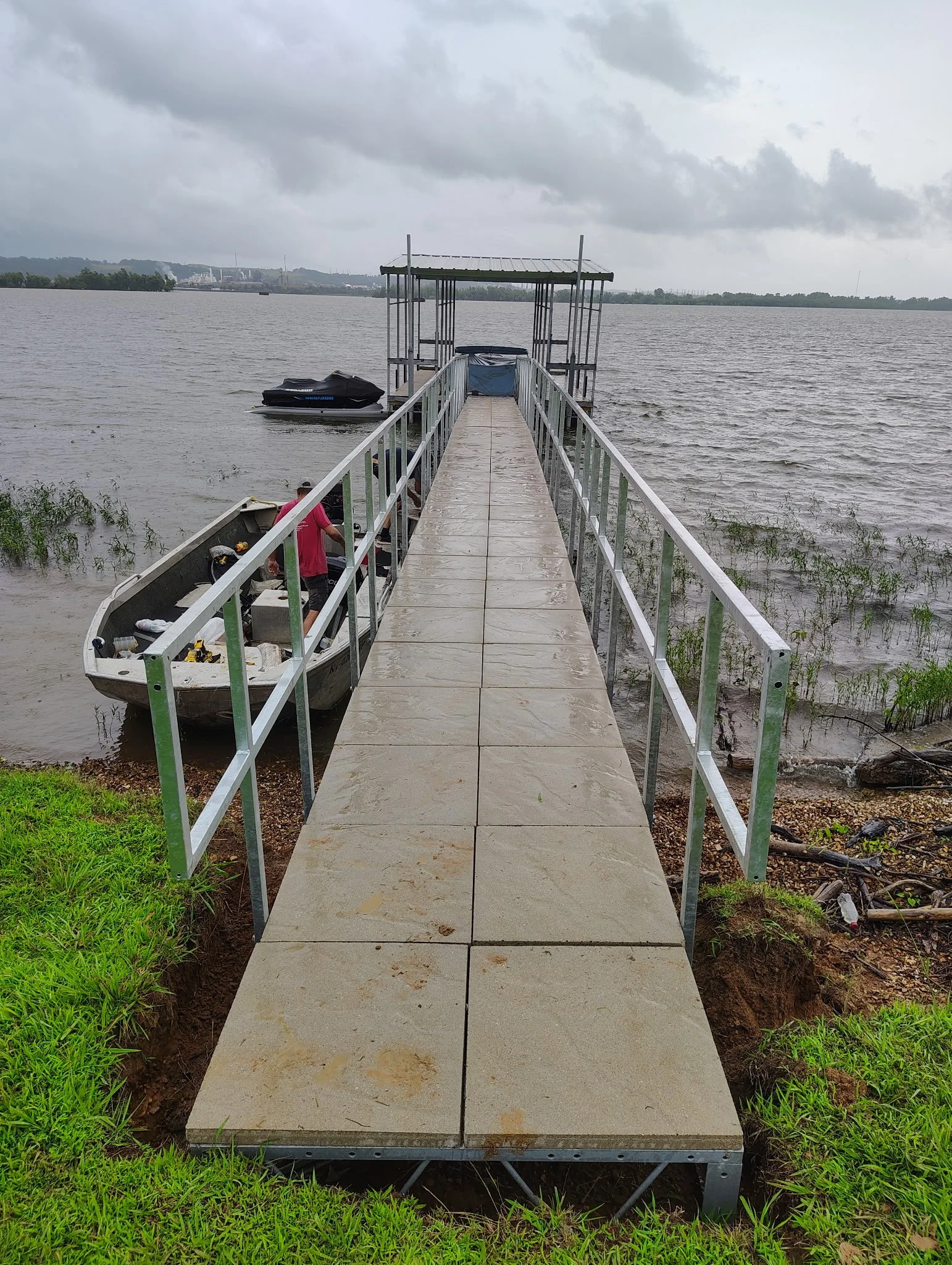 A metal and concrete dock extending into a cloudy body of water, with a boat and personal watercraft nearby, and green grass on the shore.