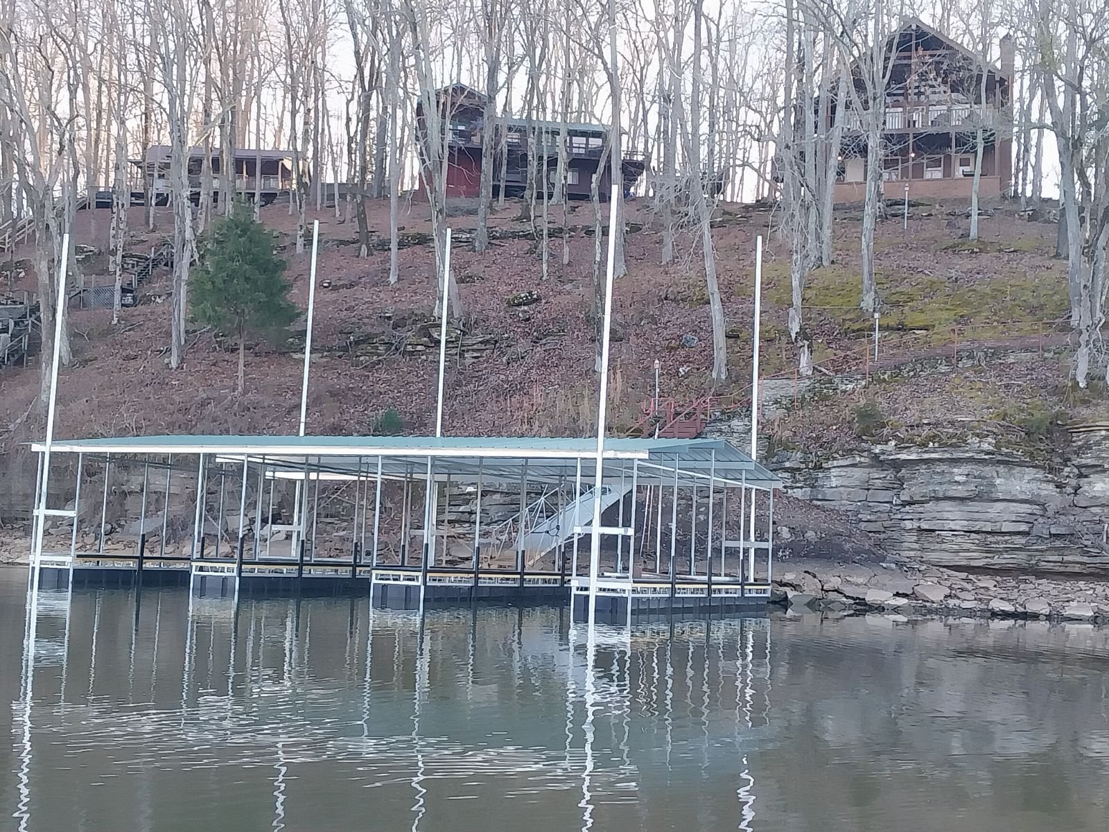 A lakeside scene with a metal boat dock extending into the water, situated at the base of a wooded hillside with houses among leafless trees.