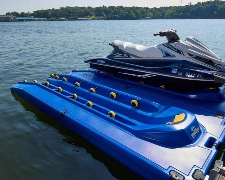 A blue jet ski ramp with yellow bumpers floating on a lake next to a white and black jet ski, with trees and sky in the background.