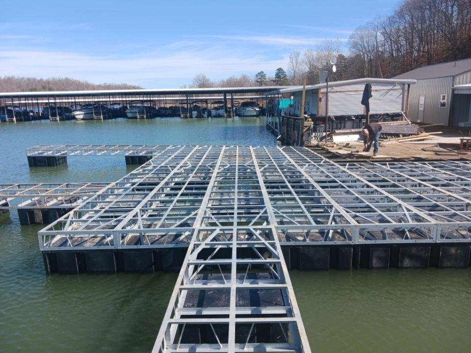 Metal framework being constructed over a lake at a marina, with boats and a covered dock in the background.