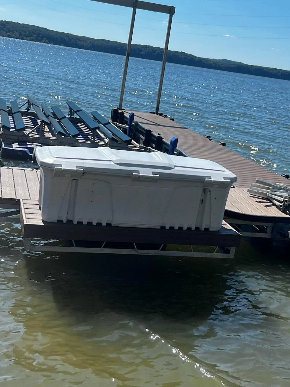 Dock on a lake with a large white storage container, and a loaded bench, with a distant tree-lined shoreline under a clear blue sky.