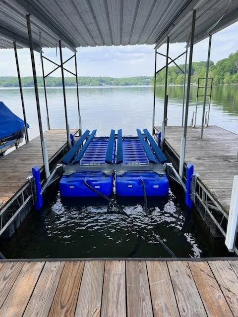 Empty boat hoist docked at a lakeside boathouse with water and green trees in the background.