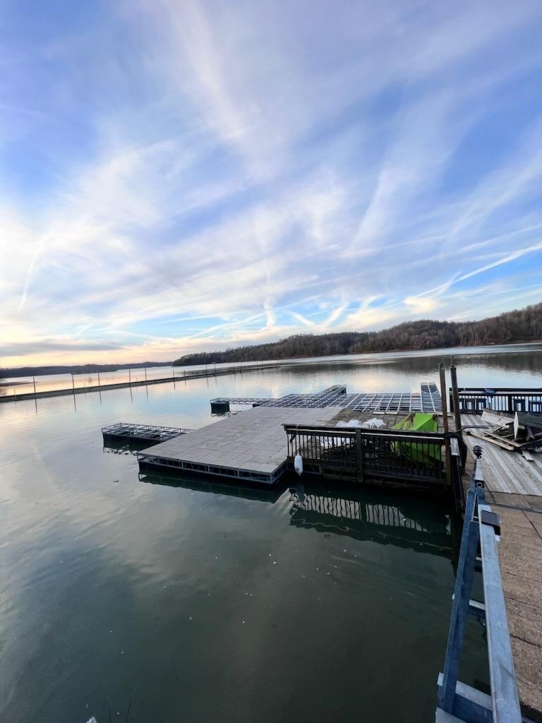 A calm lake with a waterfront dock extending into the water, surrounded by hilly terrain under a partly cloudy sky during late afternoon or early evening.