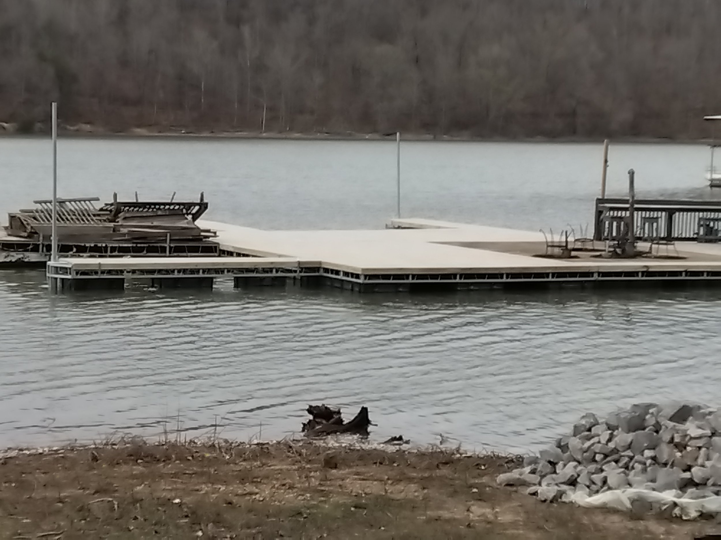 Empty dock on a lake with stacked boats and chairs, calm water, and a forested shoreline in the background.