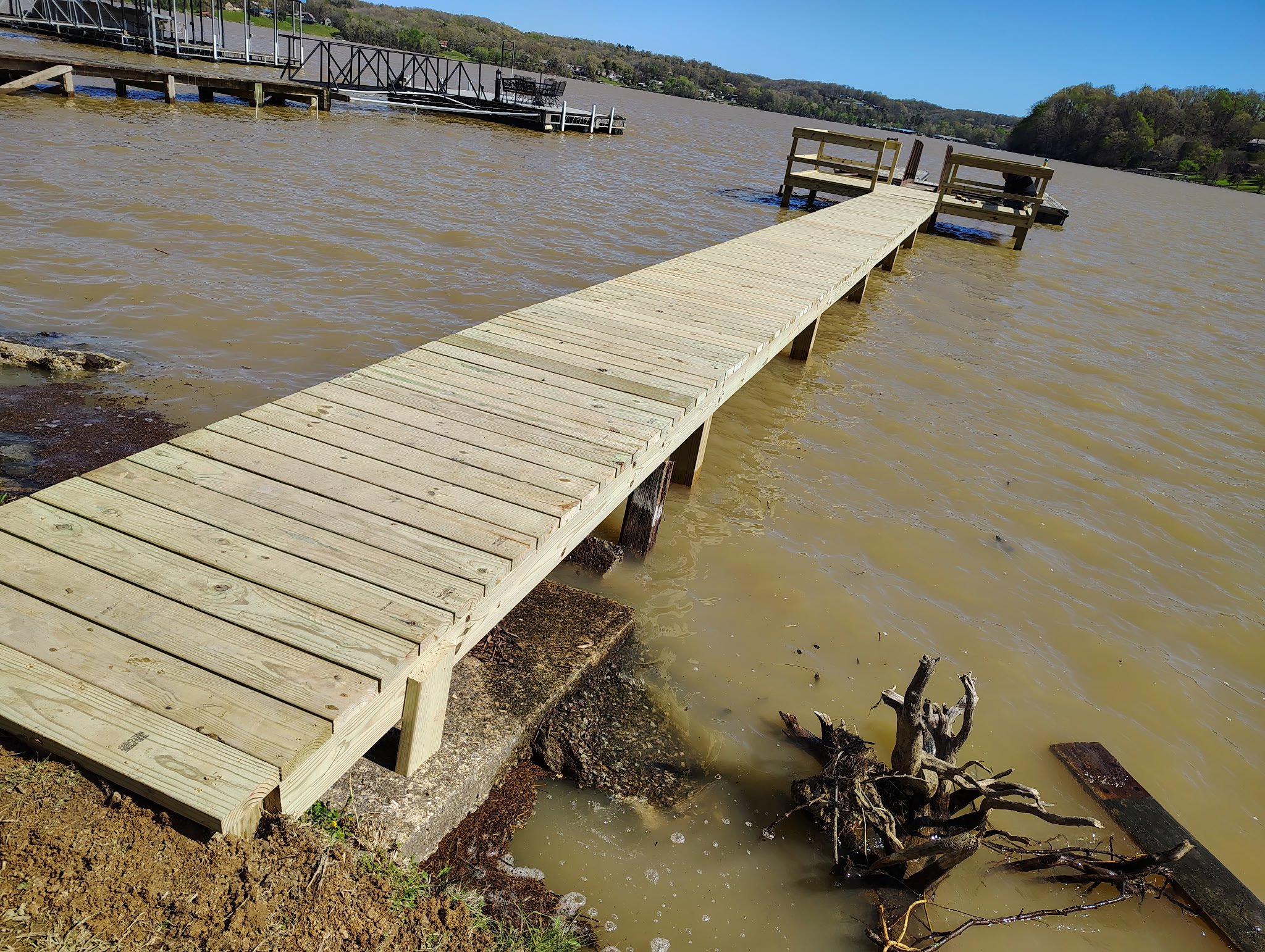 A wooden pier extending into a body of water with muddy brown color. There are small rocks and a fallen tree trunk at the shoreline. In the background, there are other docks and a landscape of trees and hills.