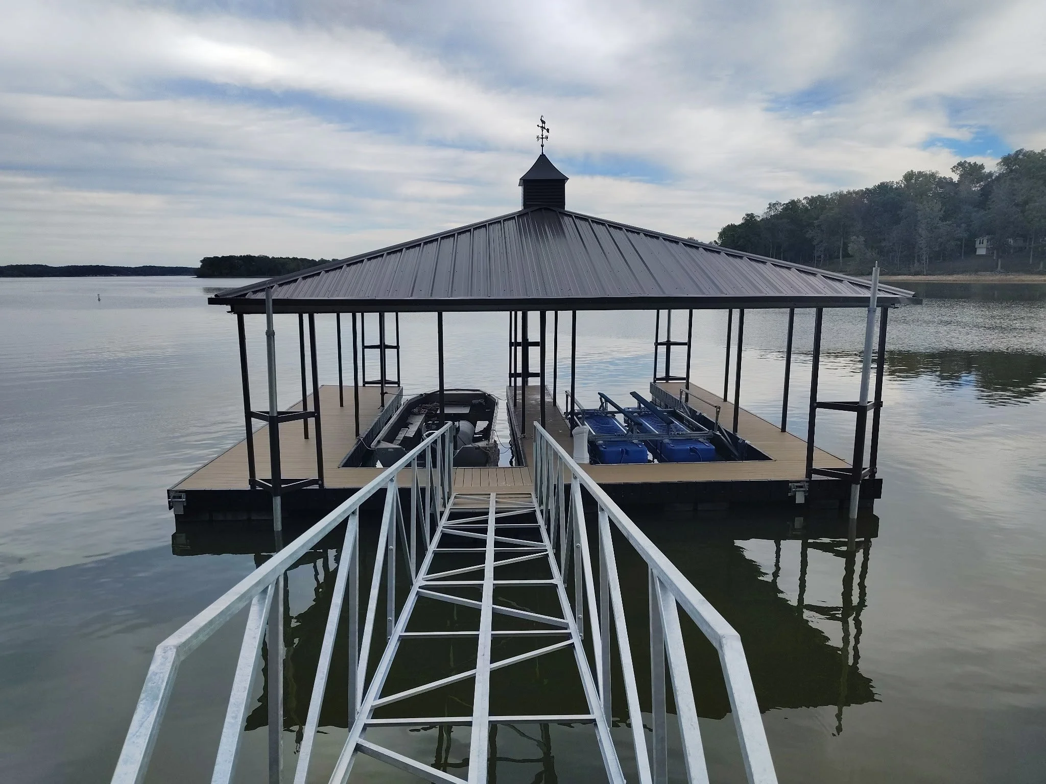 A dock leading to a covered boat house on a calm lake with trees in the background.
