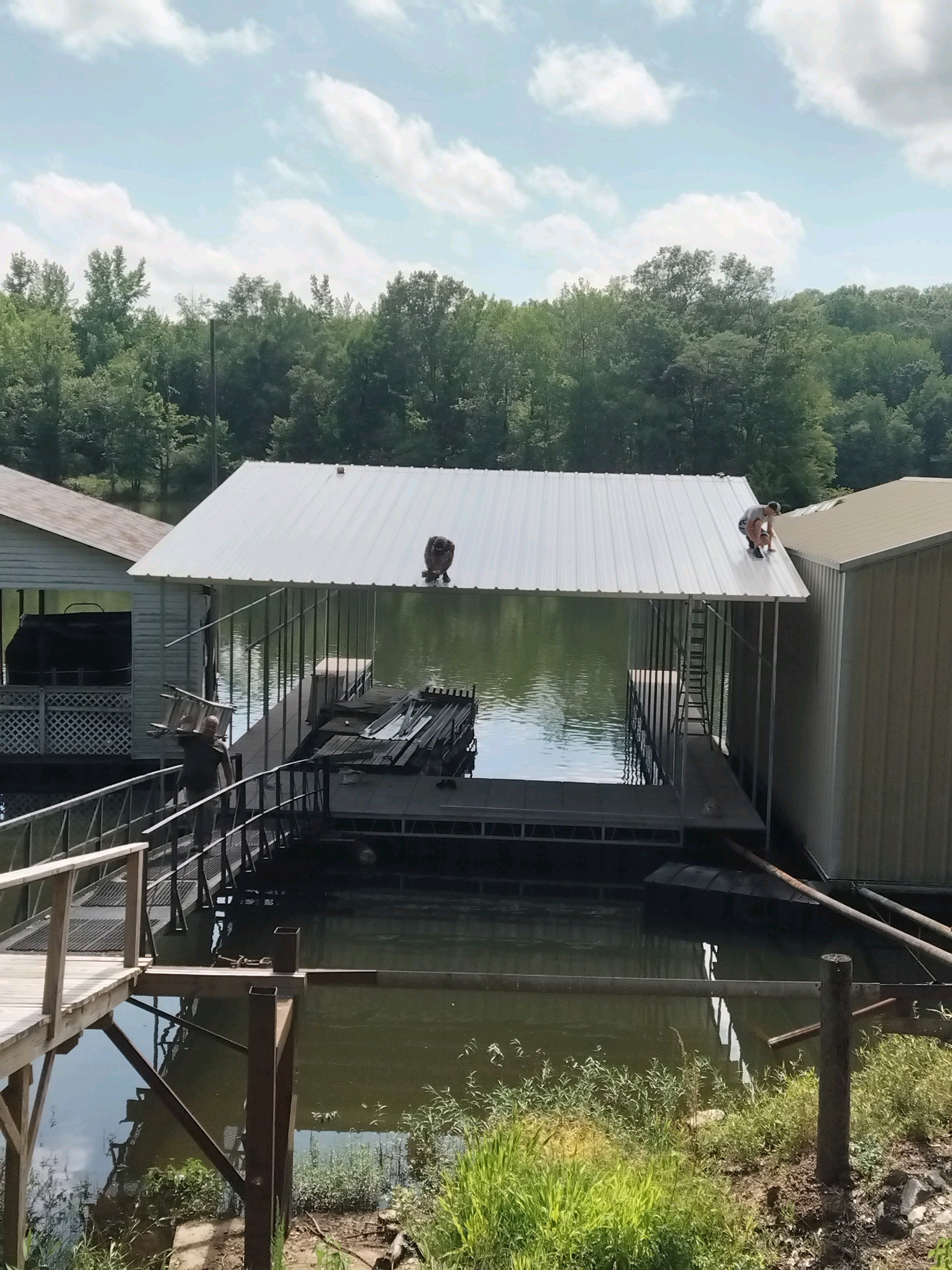 Two workers are installing a white metal roof on a boat dock in a backyard by a lake, with trees and a partly cloudy sky in the background.