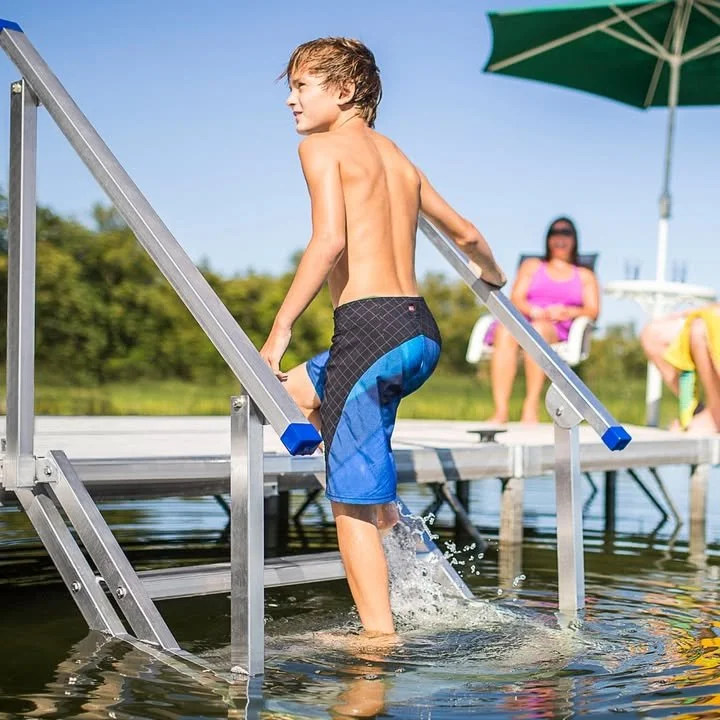 A boy in blue and black swim trunks climbing out of a lake on a metal dock with a ladder, while a woman in a pink swimsuit and sunglasses relaxes on a lounge chair under a green umbrella.