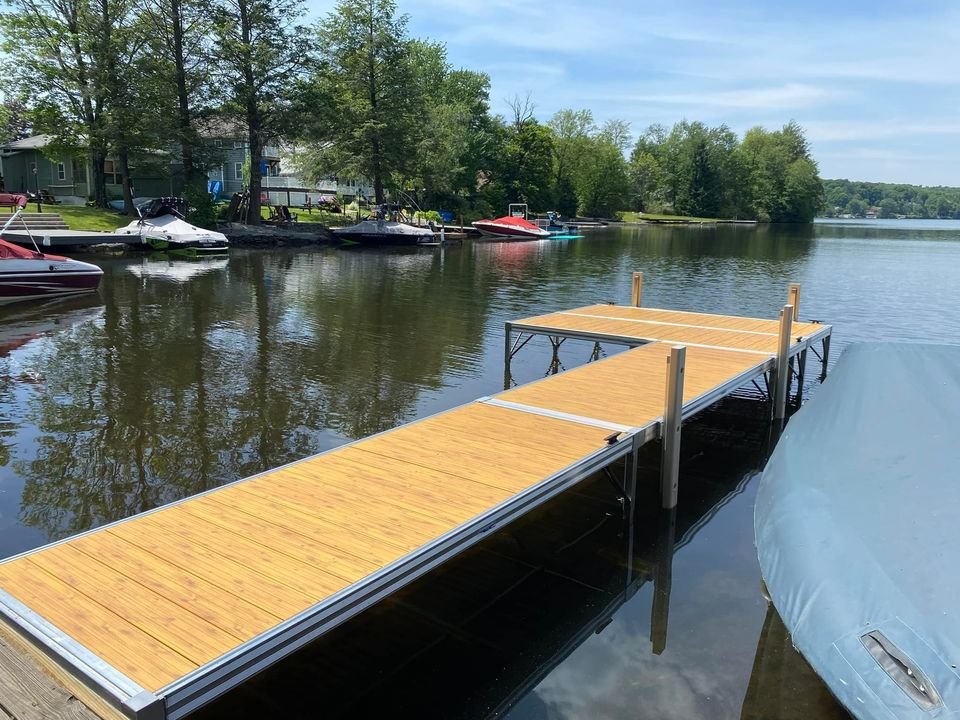 A wooden dock extending into a calm lake, with several boats moored along the shoreline, and trees in the background under a blue sky.