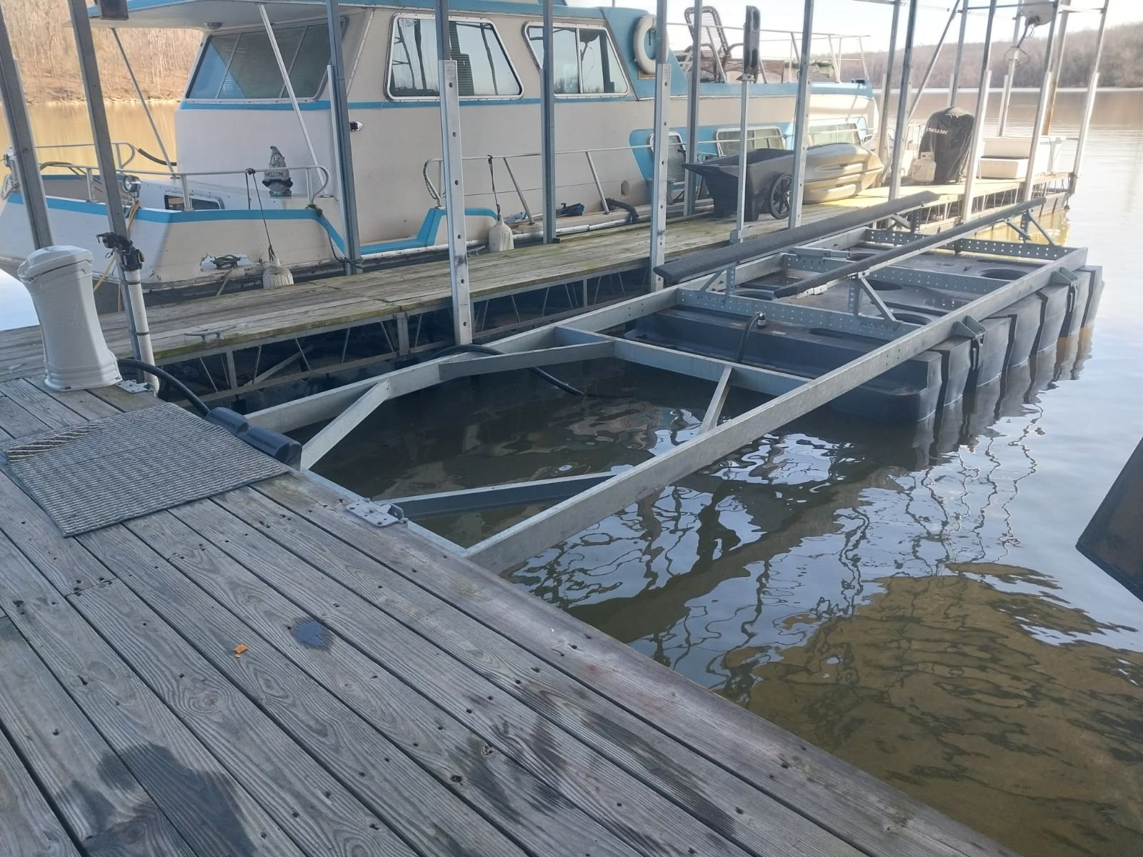 A wooden dock with metal boat lift structure in a body of water, with boats and a pontoon boat in the background.